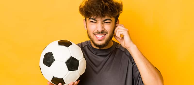 Homem jovem fazendo careta e com bola de futebol na mão